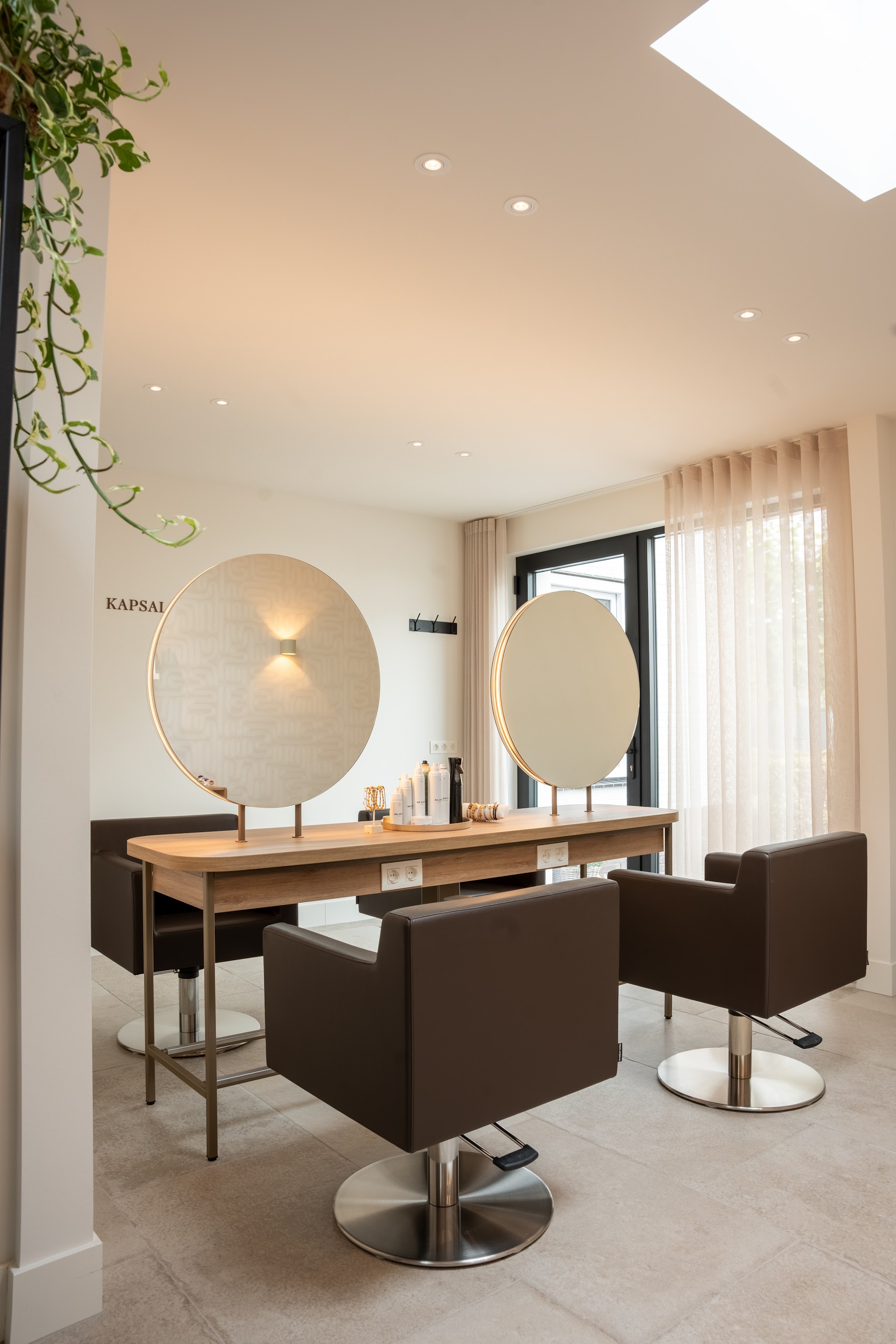 Interieur van de salon: twee bruine kappersstoelen, ronde spiegels en een houten werkblad onder een lichtkoepel